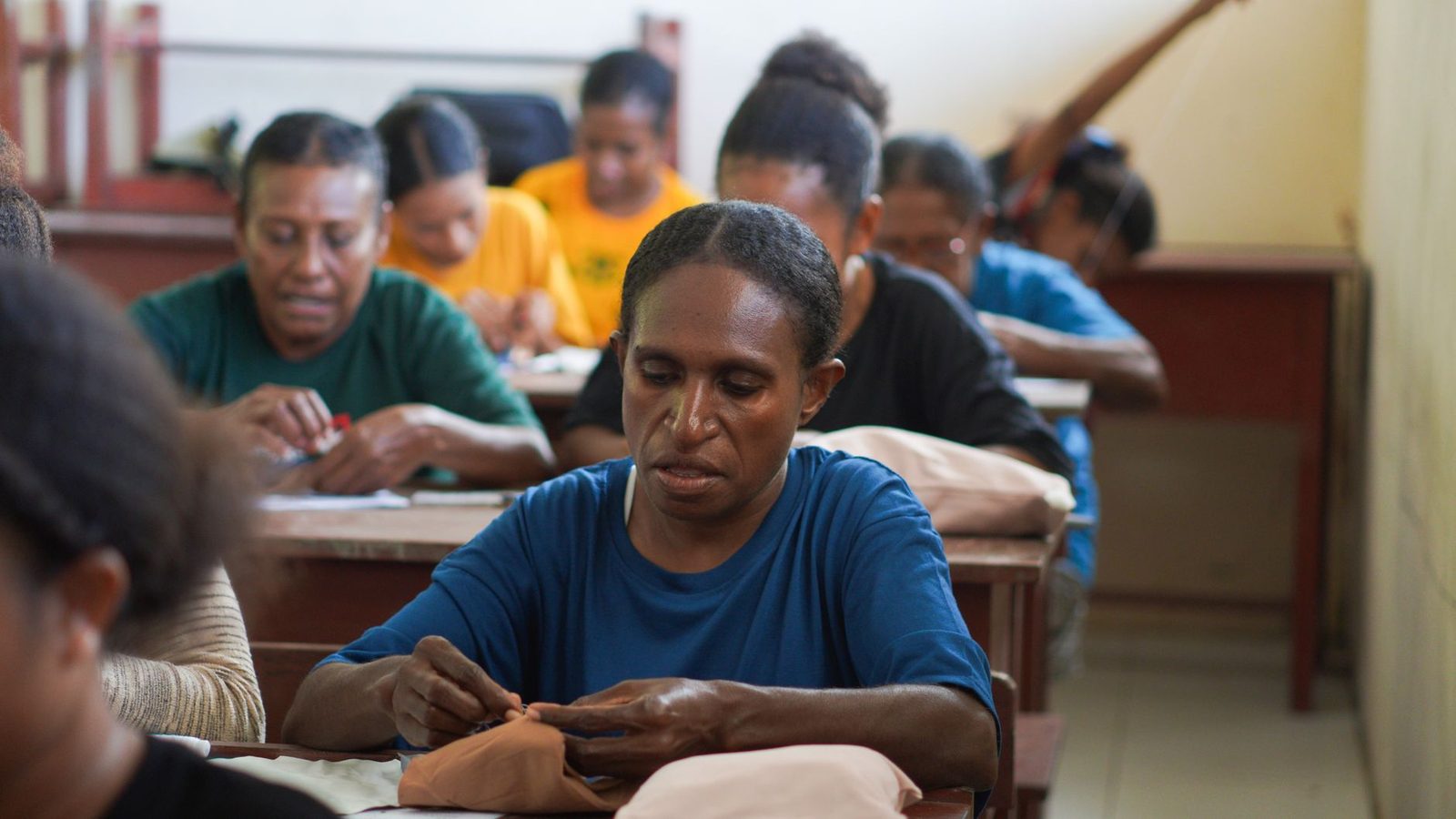 Adultos sentados a mesas durante uma formação profissional, a coser tecido à mão numa sala de aula