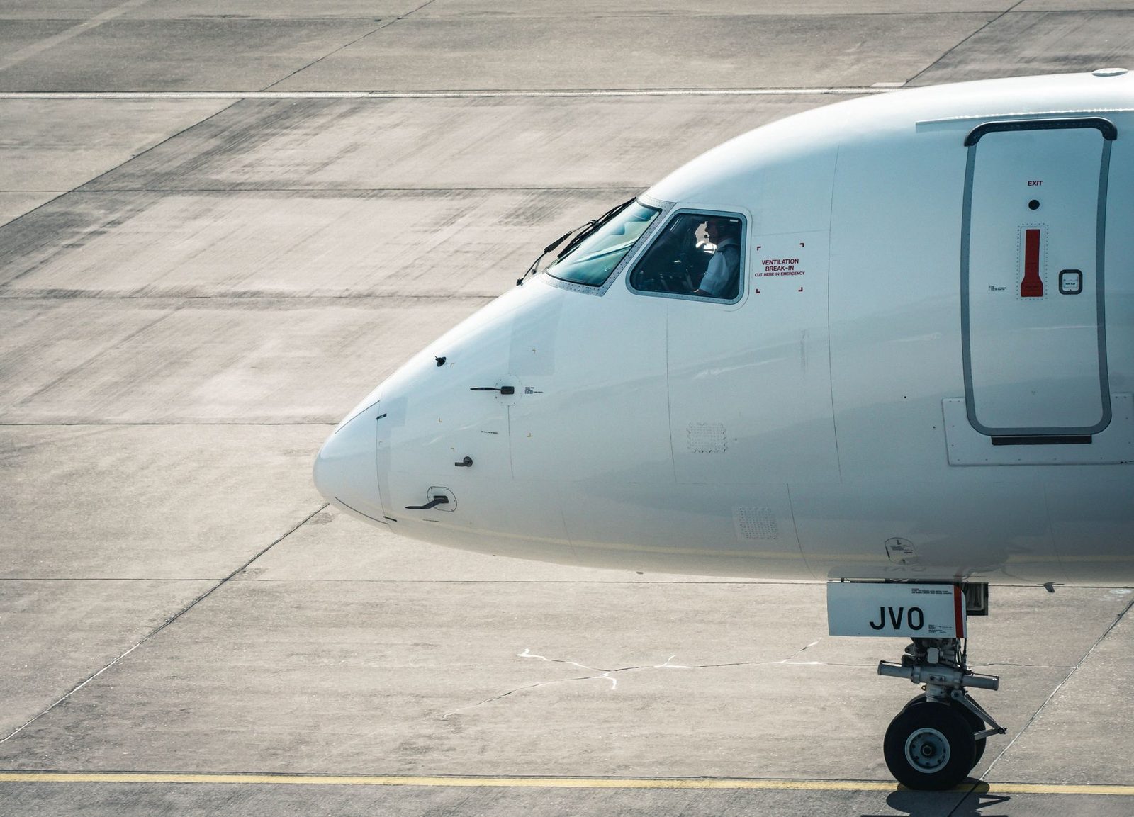 Dziób białego samolotu pasażerskiego z pilotem w kokpicie, kołującego po płycie lotniska.