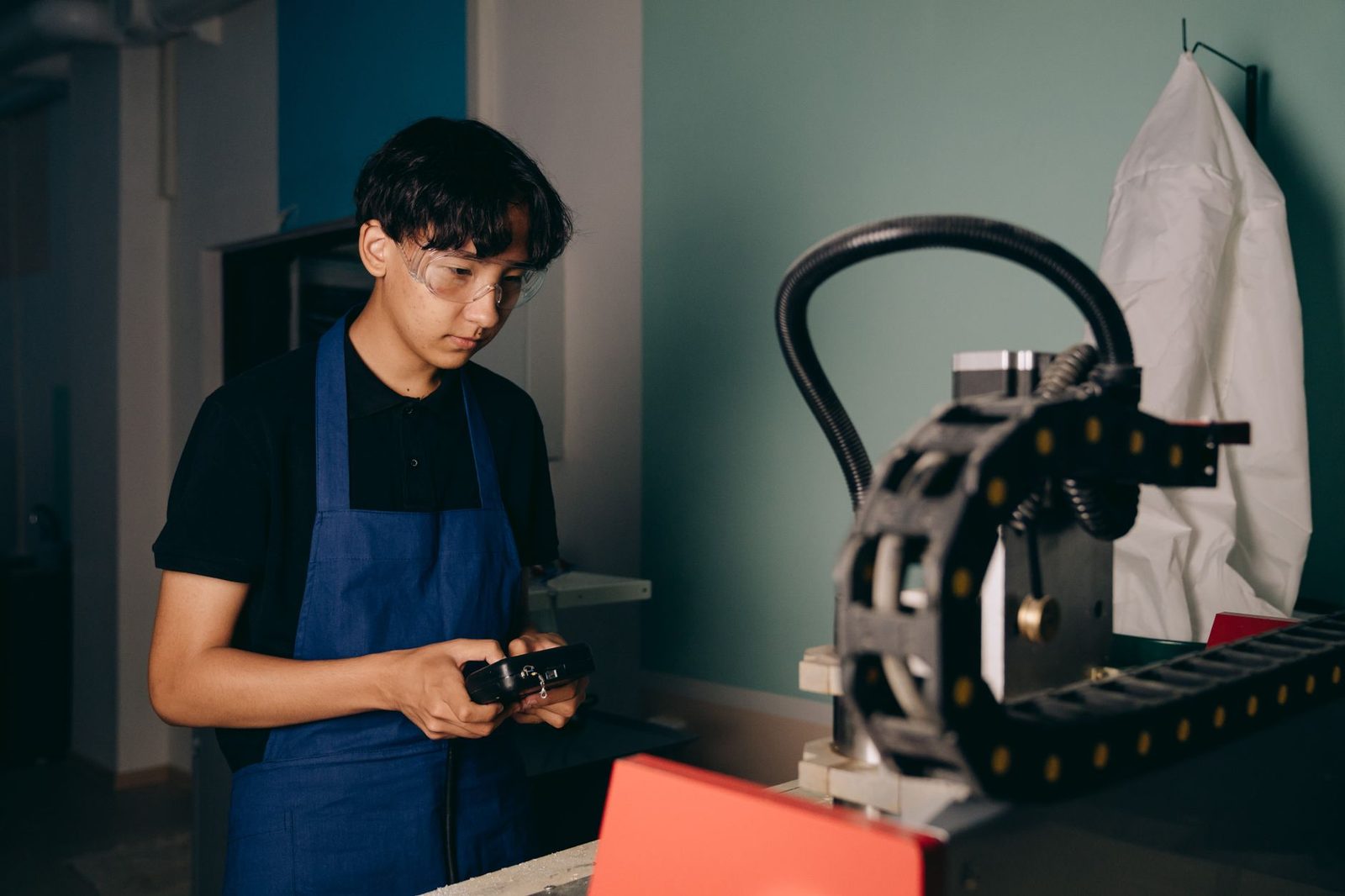 Jeune homme en tablier bleu et lunettes de protection utilisant une télécommande près d'une machine d'atelier