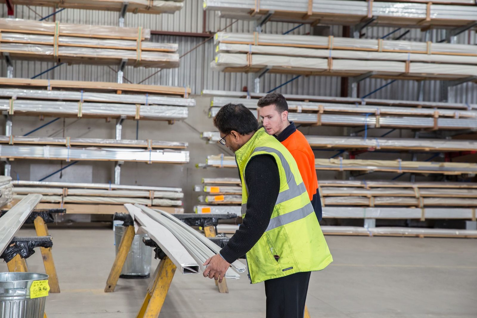 Un jeune homme en gilet orange observe un ouvrier en gilet jaune manipulant des profilés dans un entrepôt
