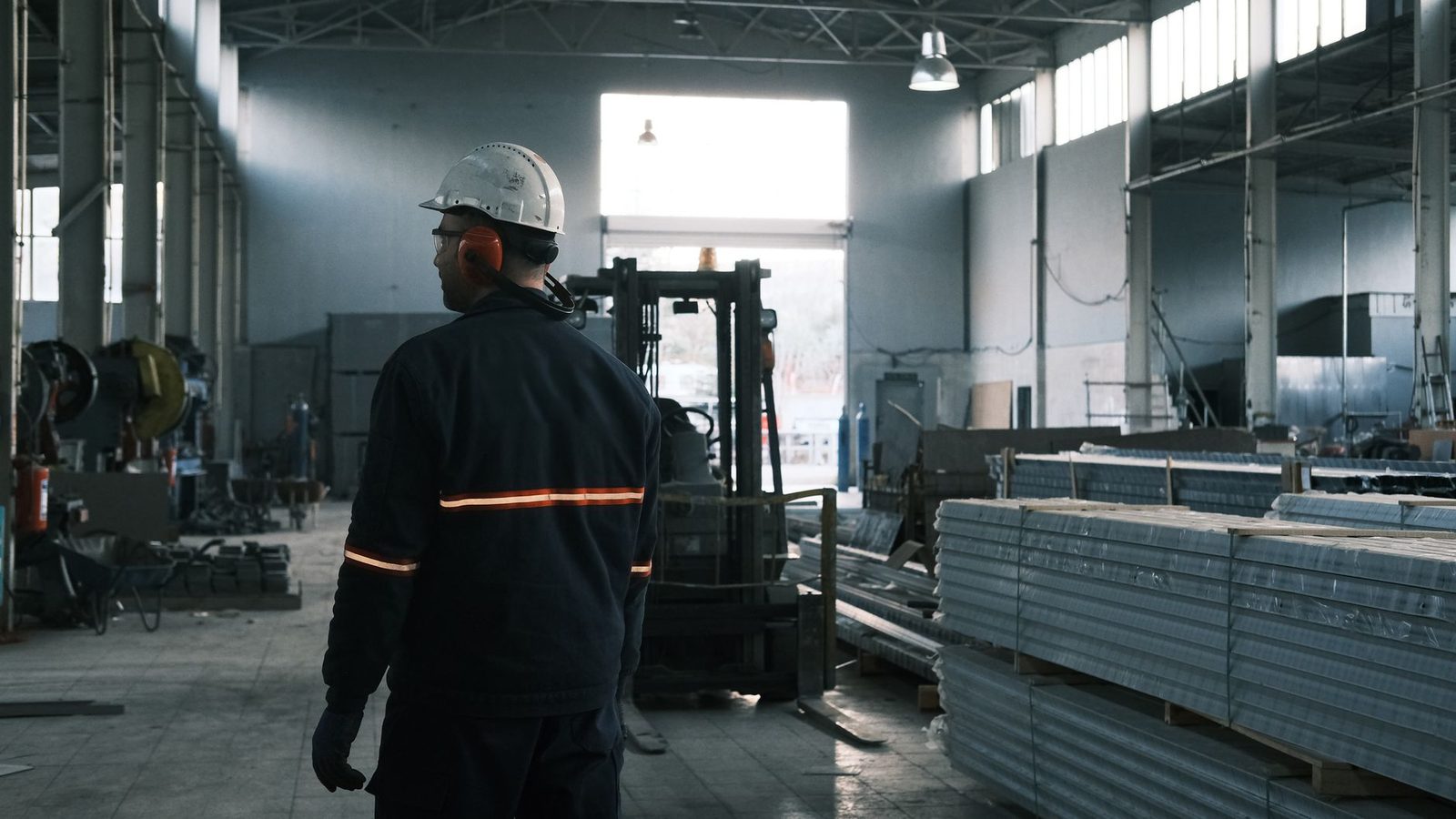 Ouvrier de dos avec casque blanc et casque antibruit dans un atelier industriel, près d'un chariot élévateur