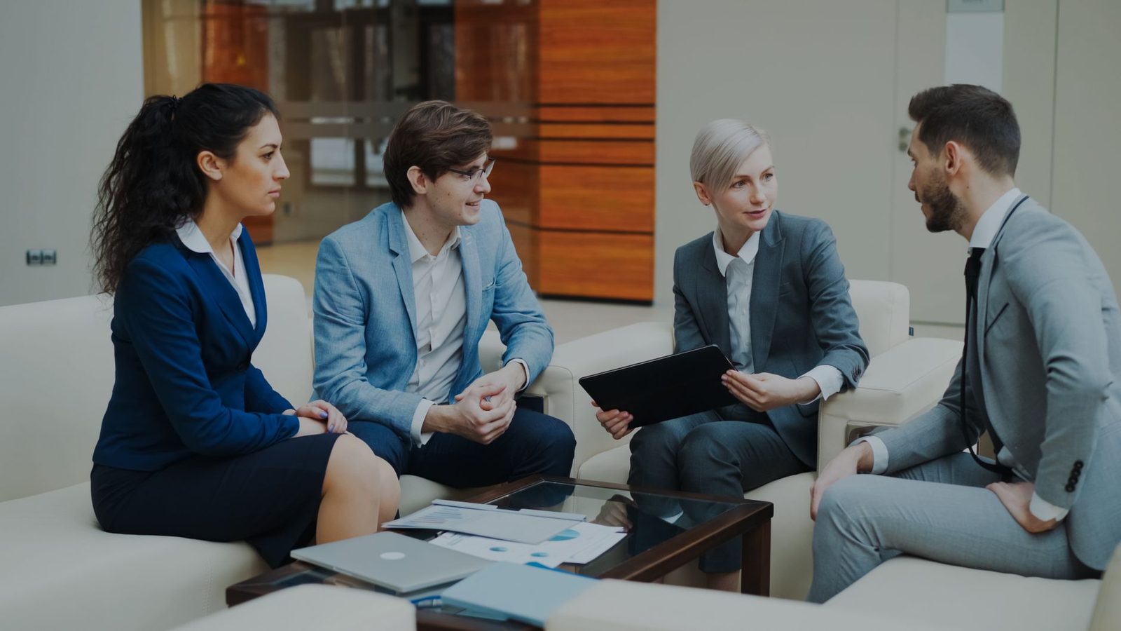 Quatre jeunes professionnels en tenue de ville discutent assis dans un salon de bureau, tablette et documents