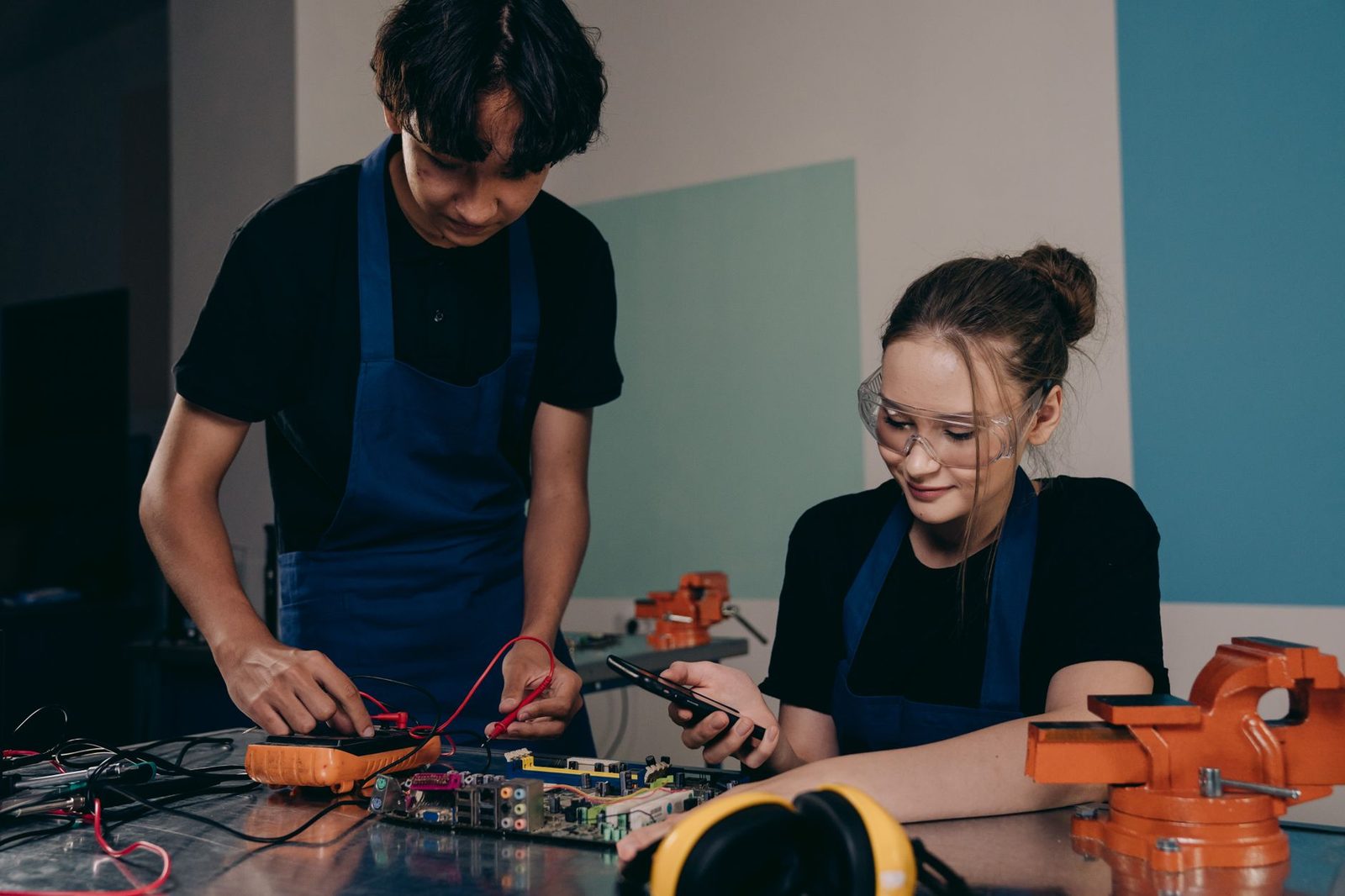 Deux jeunes en tablier bleu et lunettes de protection testent une carte électronique dans un atelier