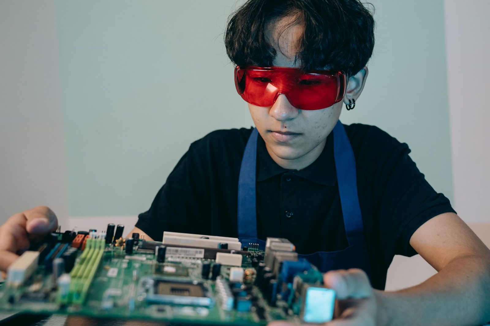 Jeune homme en polo noir et tablier, lunettes de protection rouges, travaillant sur une carte électronique