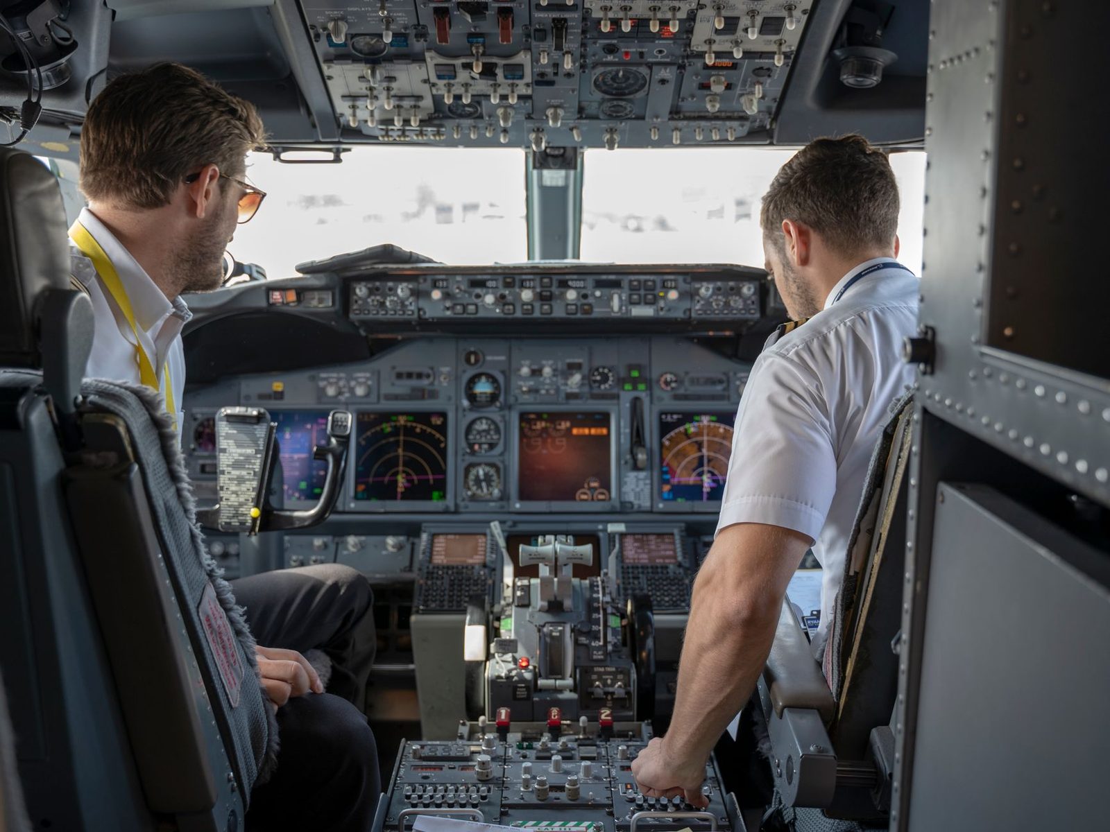 Dwóch pilotów w białych koszulach w kokpicie samolotu pasażerskiego, przed tablicą przyrządów