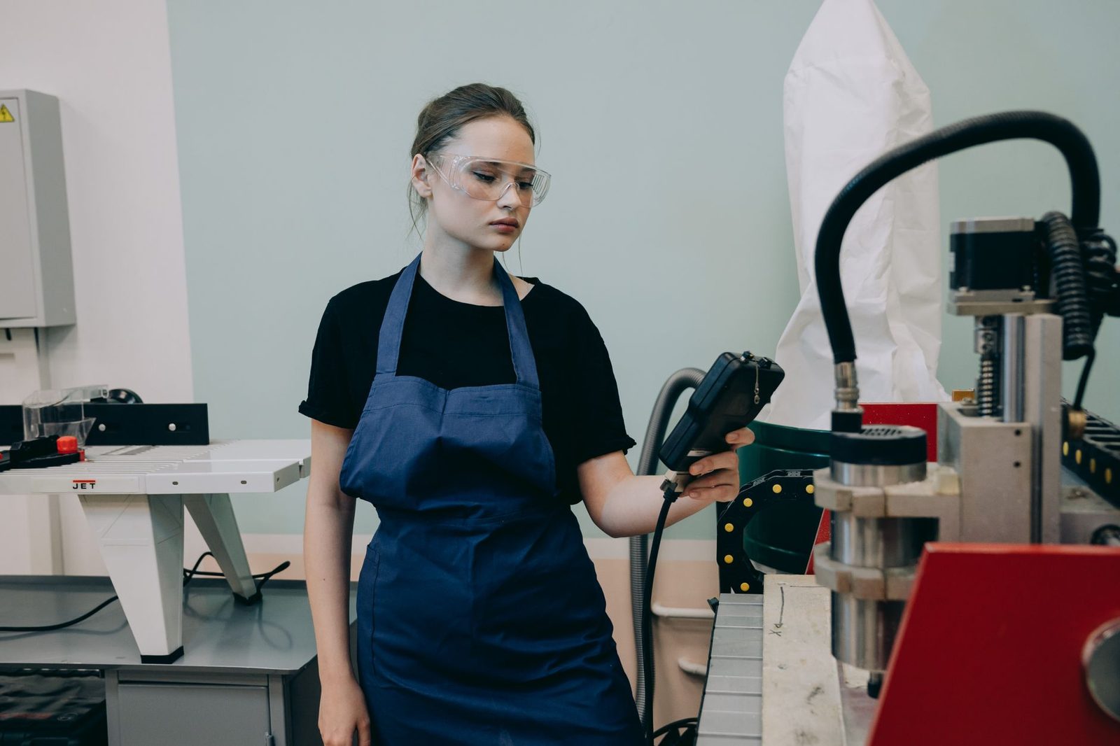 Junge Frau mit blauer Schürze und Schutzbrille bedient eine Maschine in einer Werkstatt