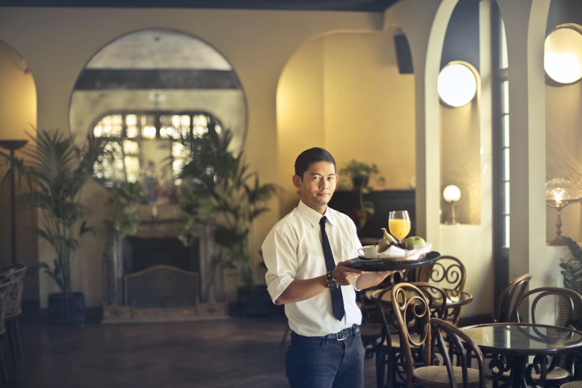 Młody kelner w eleganckim stroju niosący tacę z daniami i napojami w restauracji, ilustrujący zawody obsługi sali w systemie dualnym