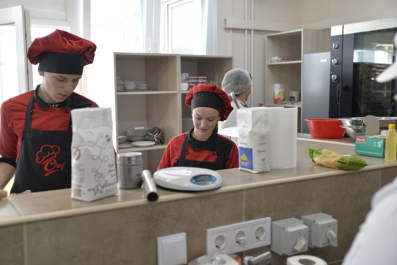 Uczniowie kucharze w białych strojach ćwiczący w kuchni szkoleniowej, ilustrujący naukę dualną w hotelarstwie i gastronomii