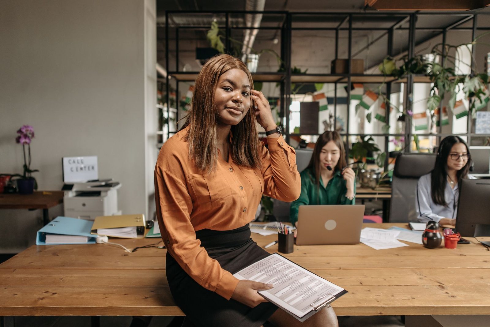 Jeune femme en chemise orange tenant un document, assise sur un bureau dans un open space avec deux collègues