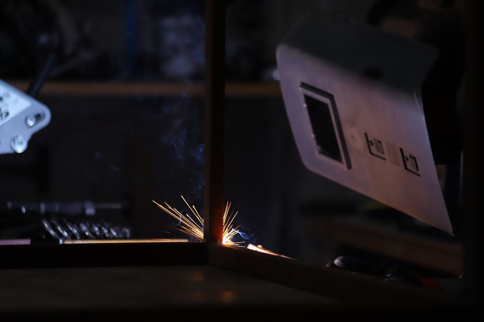 Fasci di scintille durante la saldatura di un telaio metallico in un'officina buia, maschera da saldatore accanto