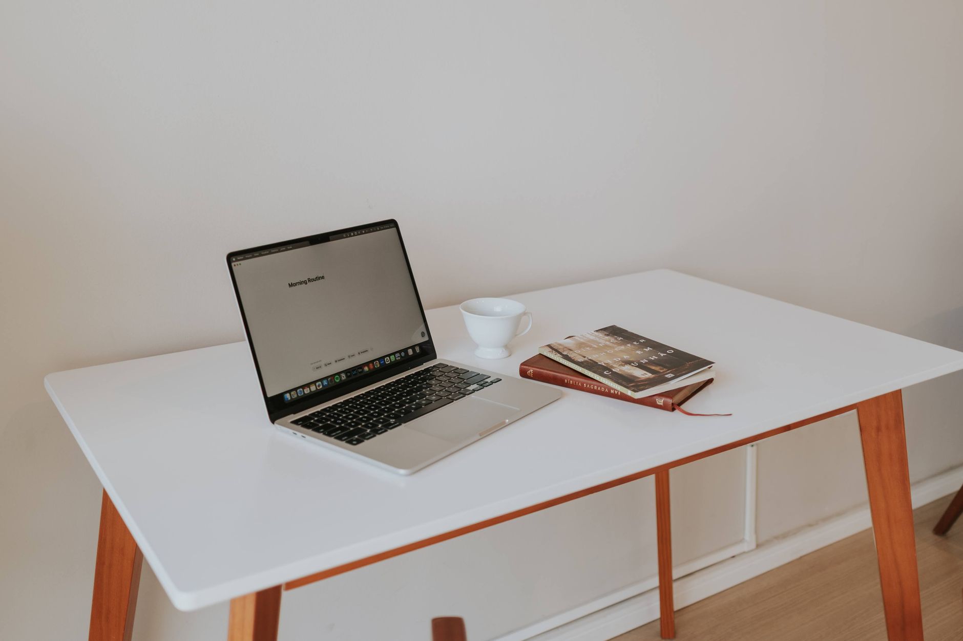 Een open laptop, een witte kop en een notitieboek op een minimalistisch wit bureau.