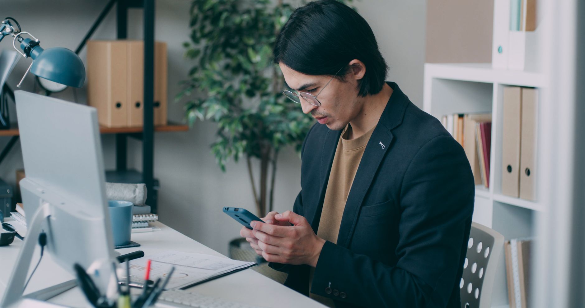 Um homem de blazer olha para o seu smartphone em frente a um computador num escritório.