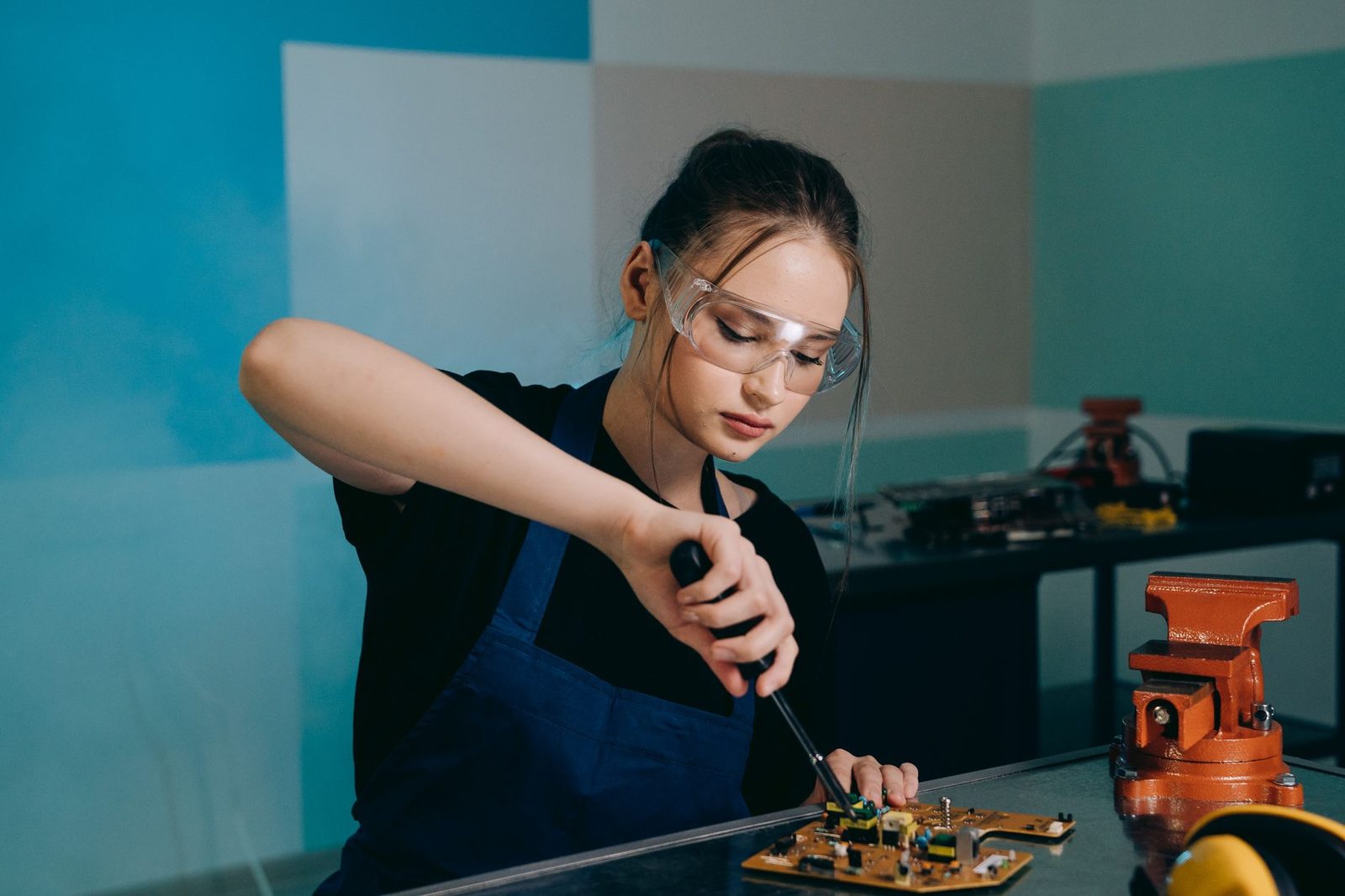 Jovem de avental azul e óculos de proteção a reparar uma placa eletrónica numa oficina