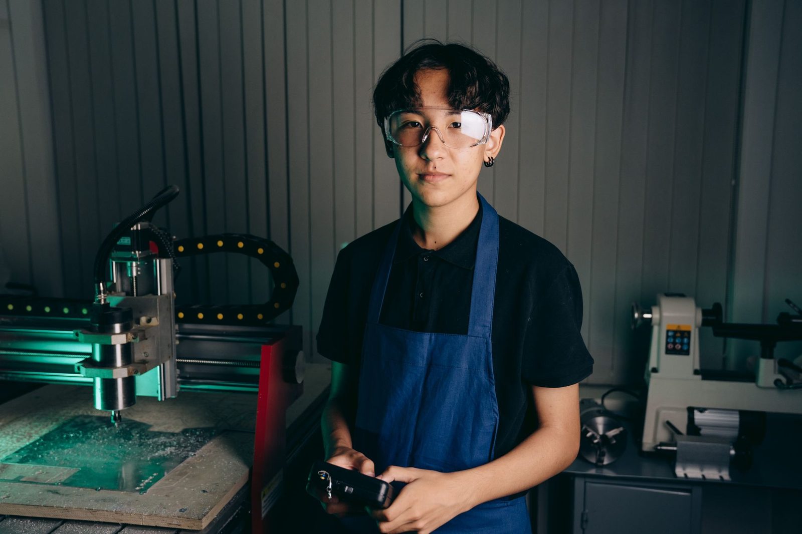Jovem aprendiz de avental azul e óculos de proteção numa oficina equipada com máquinas-ferramenta