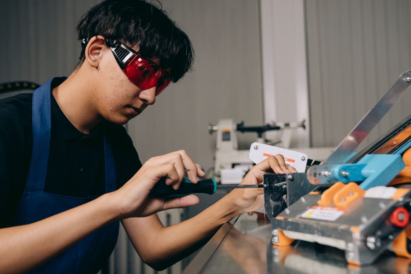 Jovem de avental e óculos de proteção vermelhos usando uma chave de fenda numa máquina numa oficina