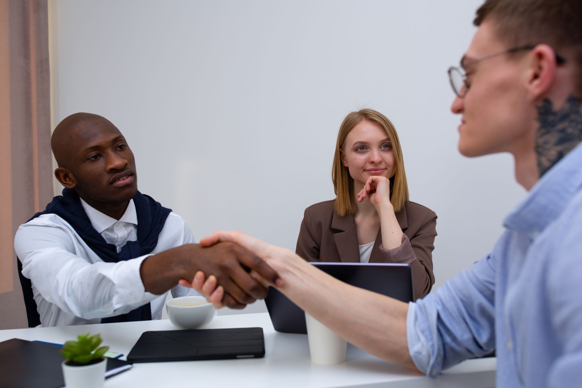 Twee mannen schudden elkaar de hand aan een bureautafel, terwijl een vrouw op de achtergrond toekijkt.