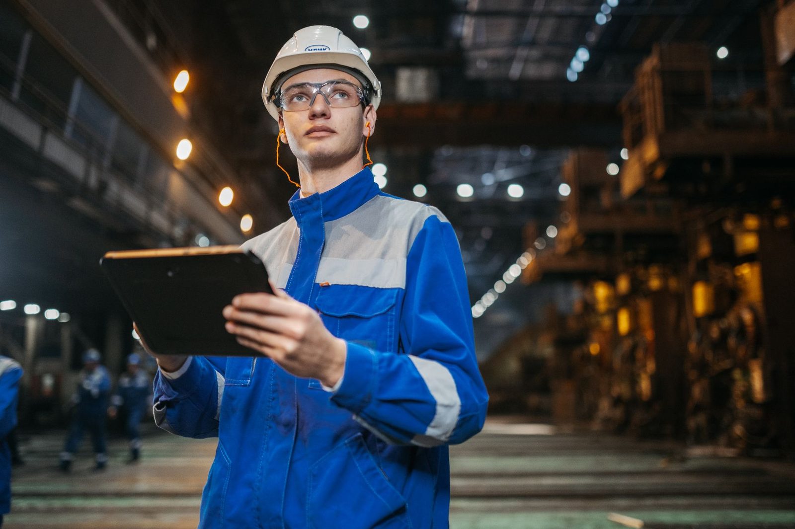 Joven técnico con mono azul y casco blanco sosteniendo una tableta en un taller industrial