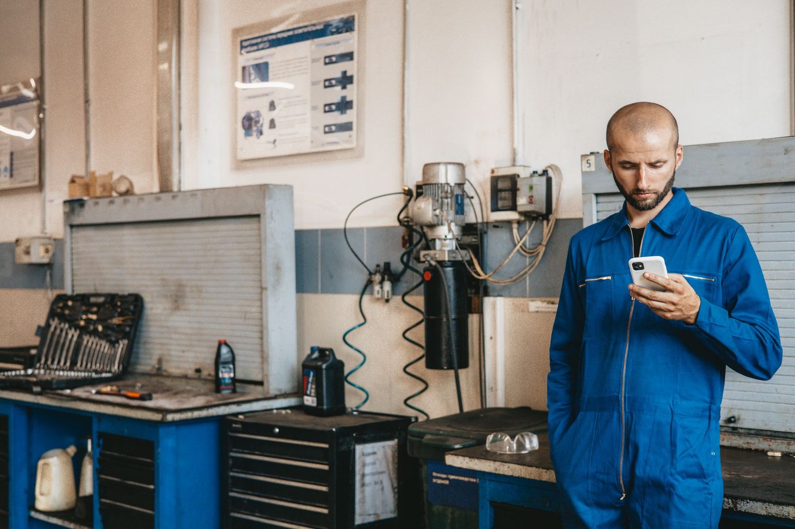 Young man in blue overalls checking his smartphone in a mechanic's workshop fitted out with tools